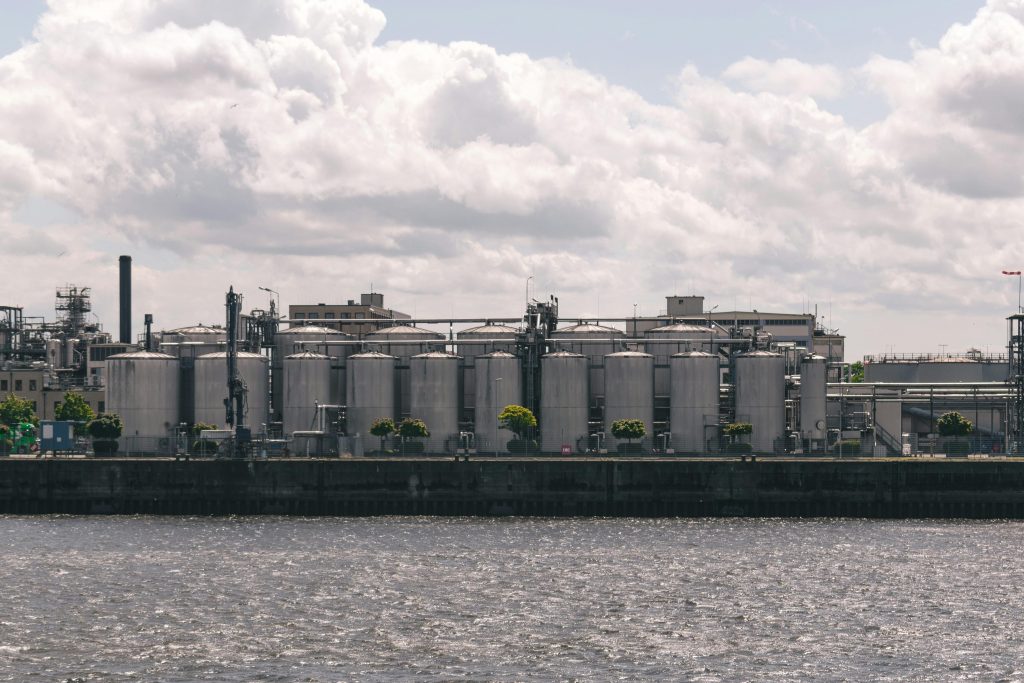 A riverside view of industrial silos and factories under cloudy skies.